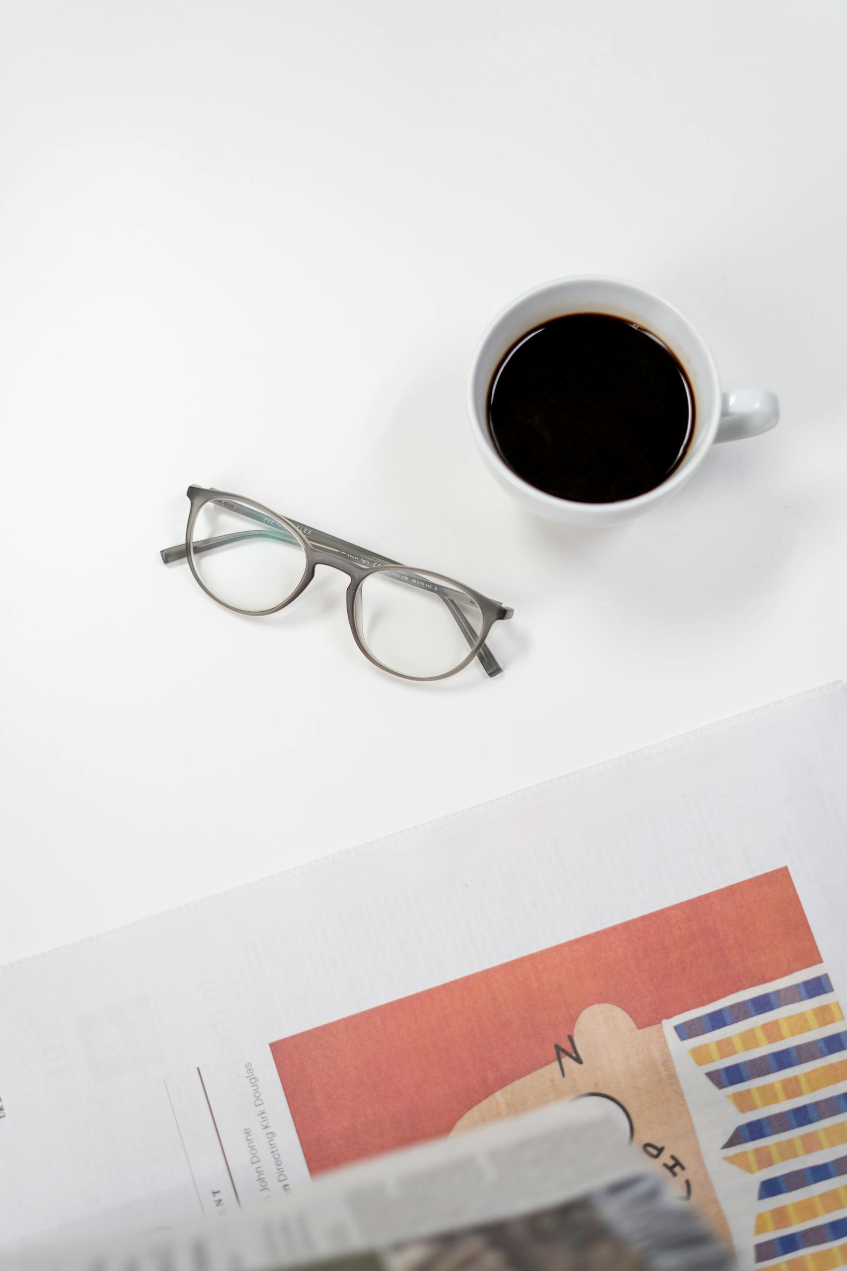 Flat lay of coffee, glasses, and newspaper on a white desk, perfect for a home office setup.